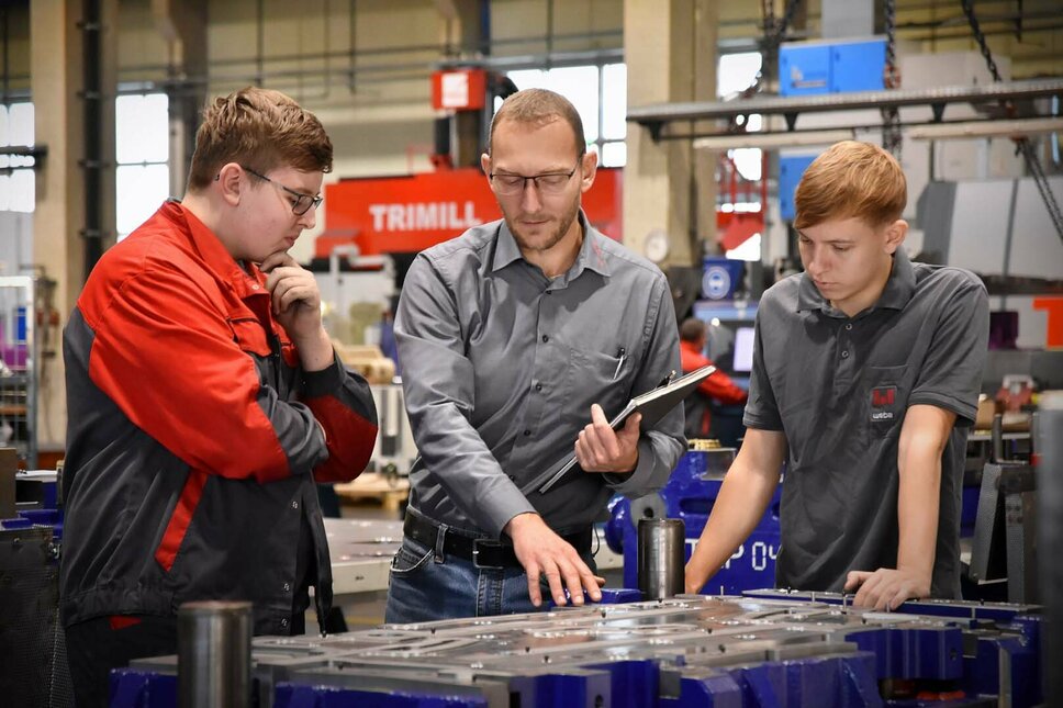 Das Bild zeigt den Lehrlingsausbilder, der zwei Lehrlingen in der Werkzeugbautechnik in einer Produktionsumgebung ein Stanz- und Umformwerkzeug erklärt. Er zeigt auf bestimmte Teile des Werkzeugs und erklärt die Funktionen und den Zweck jeder Komponente. Die Lehrlinge hören aufmerksam zu und stellen Fragen, während sie lernen. Die Szene vermittelt den Eindruck von Wissensaustausch und praktischer Schulung in einem professionellen Arbeitsumfeld.