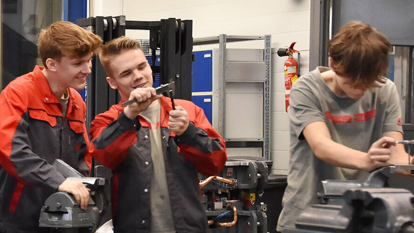 Foto von Lehrlingen in einer Lehrwerkstätte in ihrer Lehrstelle in Steyr bei der Arbeit: Zwei Lehrlinge unterhalten sich über eine technische Zeichnung, zwei weitere messen ein Stahlstück und ein Lehrling arbeitet an einem Spannbock.