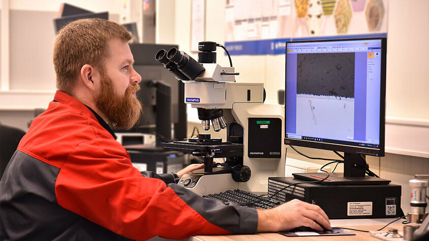 Ein Mitarbeiter der Qualitätssicherung sitzt an einem Schreibtisch und blickt konzentriert auf einen Computerbildschirm, auf dem Ergebnisse der Materialprüfung angezeigt werden. Im Hintergrund des Raumes ist ein Mikroskop erkennbar, das auf die Wichtigkeit genauer Untersuchungen in diesem Bereich hinweist. 