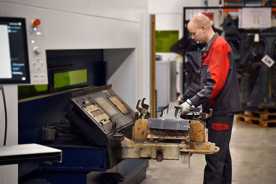 Ein Mitarbeiter bedient eine Laserschneidmaschine in einer Produktionshalle. Er hält ein Werkstück aus Metall, das gerade in die Maschine zum Laserschneiden eingesetzt wird. Die Arbeitsumgebung ist sauber und gut organisiert, mit verschiedenen Werkzeugen und Materialien im Hintergrund.