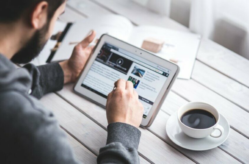 A man is working on a tablet. Coffee and an open newspaper can be seen on the table in front of him.