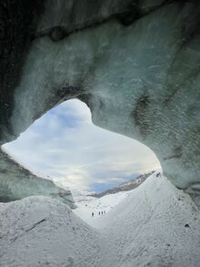 Breathtaking view from a glacier cave overlooking a snow-covered landscape with distant mountains. Three hikers are visible in the distance.