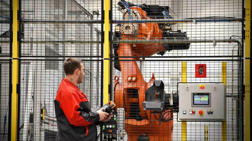 Ein Mitarbeiter in roter Arbeitskleidung bedient einen Industrieroboter in einer gesicherten Produktionszelle. Der Roboterarm ist hinter einem Sicherheitszaun sichtbar, und der Bediener hält ein Steuergerät in der Hand, um die Bewegungen des Roboters zu überwachen oder anzupassen. Neben dem Roboter befindet sich ein Bedienfeld mit einem digitalen Display, das zur Kontrolle und Überwachung der Roboterprozesse dient. Die Szene zeigt den Einsatz von Automatisierung und fortschrittlicher Robotik in der mechanischen Bearbeitung.