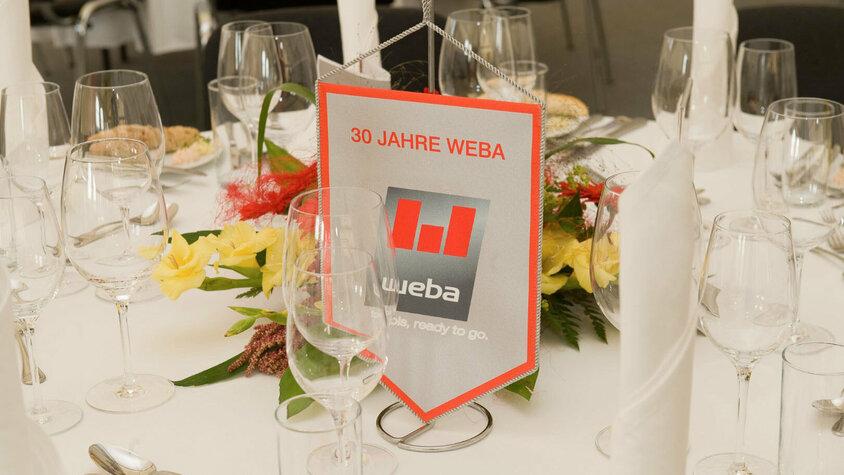 Festliche Tischdeko beim VIP-Abend des 30-Jährigen Jubiläumsfests, mit Wimpel, auf dem das weba-Logo zu sehen ist, umgeben von elegantem Dekor.