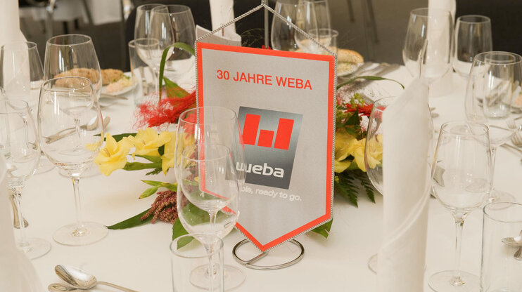 Festliche Tischdeko beim VIP-Abend des 30-Jährigen Jubiläumsfests, mit Wimpel, auf dem das weba-Logo zu sehen ist, umgeben von elegantem Dekor.