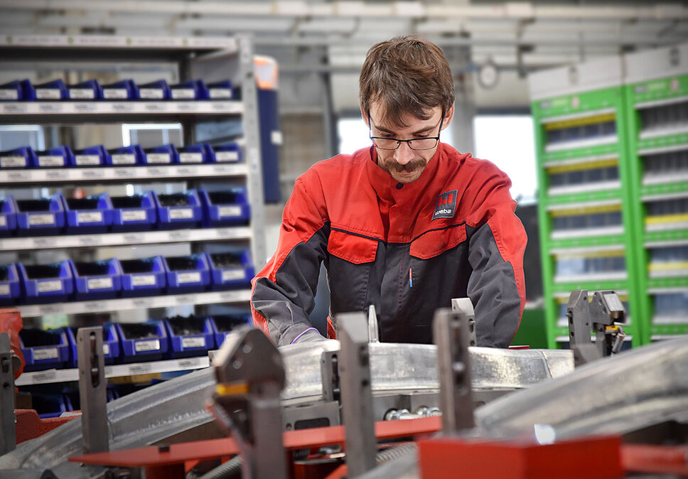 Ein Werkzeugbautechniker baut konzentriert ein Presswerkzeug für anschließenden Werkzeug-Tryout und die Umformung von Strukturbauteilen zusammen; Im Hintergrund sind unscharf Regale mit Hilfs- und Betriebsstoffen 