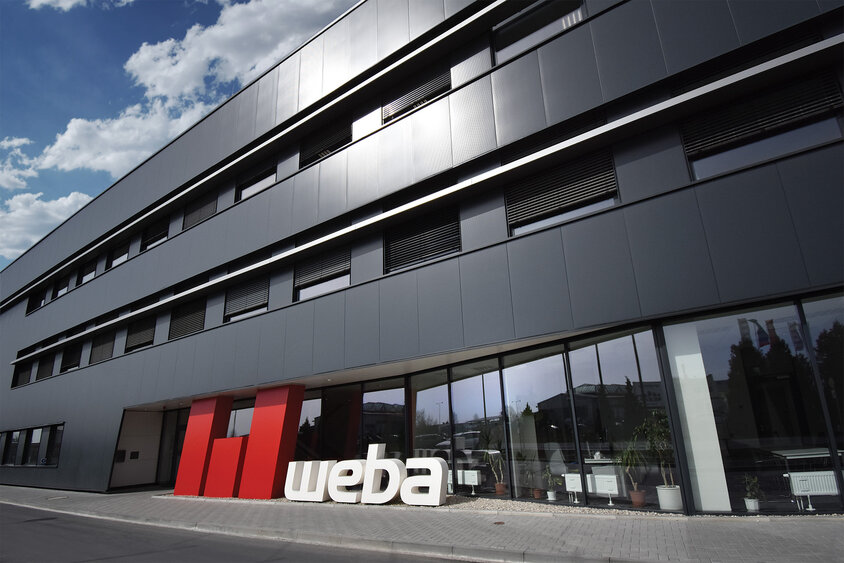 Außenaufnahme der Gebäudefront von Weba in Olomouc, charakterisiert durch eine moderne Anthrazitfassade. Vor dem Eingang ist ein großes, auffälliges 3D-Logo von WebA in Rot und Weiß platziert, das markant und einladend wirkt und die Firmenidentität stark hervorhebt.