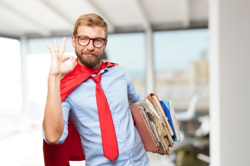 Ein Mann in Anzug mit rotem Umhang und roter Krawatte hält Akten und zeigt das OK-Zeichen, um Zustimmung und Kompetenz zu signalisieren.