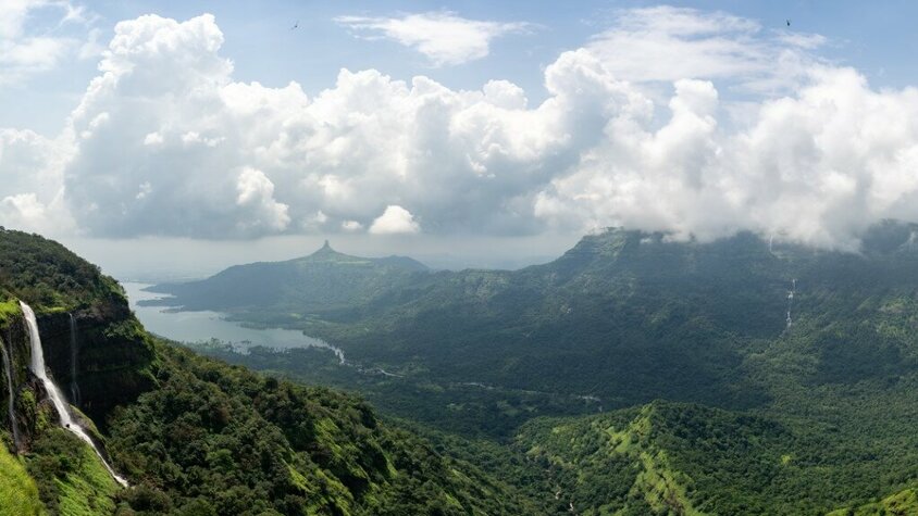 Ein malerisches Panorama mit grünen Hügeln, dichten Wäldern und Wasserfällen, umgeben von einer nebligen Berglandschaft unter einem Himmel mit weißen, dramatischen Wolken.